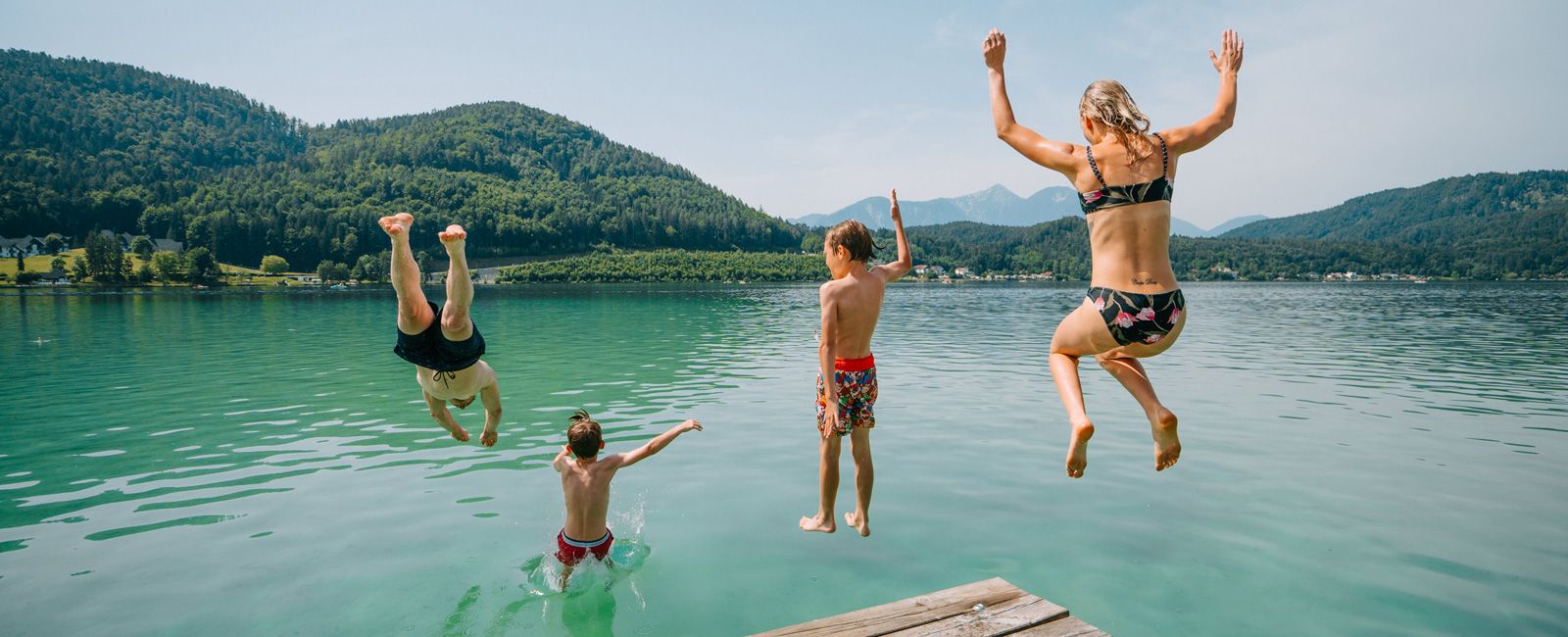Drei Kinder springen von einem Steg in den Klopeiner See, im Hintergrund bewaldete Hügel